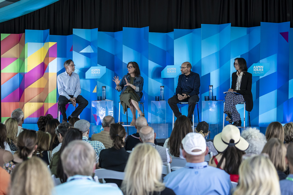 Food is Medicine panel on stage at Aspen Ideas Health 2025. They are seated on metal stools in front of a colorful backdrop. Far left, variations of wavering colors: orange, yellow, pink, green, blue, and purple. The rest of the backdrop are geometrical shapes in varying shades of blue with three prominent boxes with the title: Aspen Ideas Health in white lettering. Seated from left to right: Corby Kummer Executive Director, Food and Society Program, Aspen Institute Hilary Seligman Professor of Medicine, University of California San Francisco John Lumpkin President, Blue Cross and Blue Shield of North Carolina Foundation Wendy Slusser Associate Vice Provost, Semel Healthy Campus Initiative Center, University of California at Los Angeles