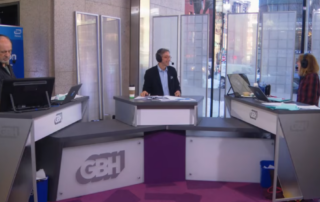 A screenshot of the Boston Public Radio set at the Boston Public Library. Behind the GBH news desks (left to right): Jim Braude (co-host), Corby Kummer (guest) and Margery Eagan (co-host) eagerly discuss current American food culture.