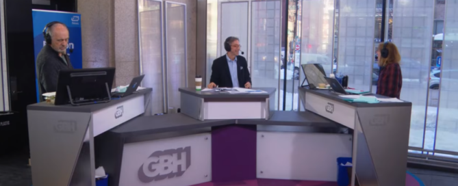 A screenshot of the Boston Public Radio set at the Boston Public Library. Behind the GBH news desks (left to right): Jim Braude (co-host), Corby Kummer (guest) and Margery Eagan (co-host) eagerly discuss current American food culture.