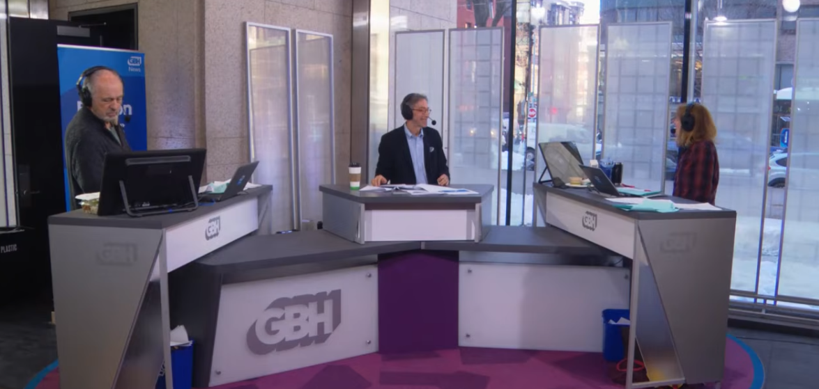 A screenshot of the Boston Public Radio set at the Boston Public Library. Behind the GBH news desks (left to right): Jim Braude (co-host), Corby Kummer (guest) and Margery Eagan (co-host) eagerly discuss current American food culture.