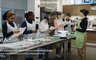 Medical students standing front of a metal workstation in a teaching kitchen. A woman in a bright green apron leads the class.