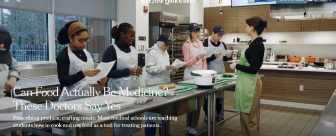 Medical students standing front of a metal workstation in a teaching kitchen. A woman in a bright green apron leads the class.