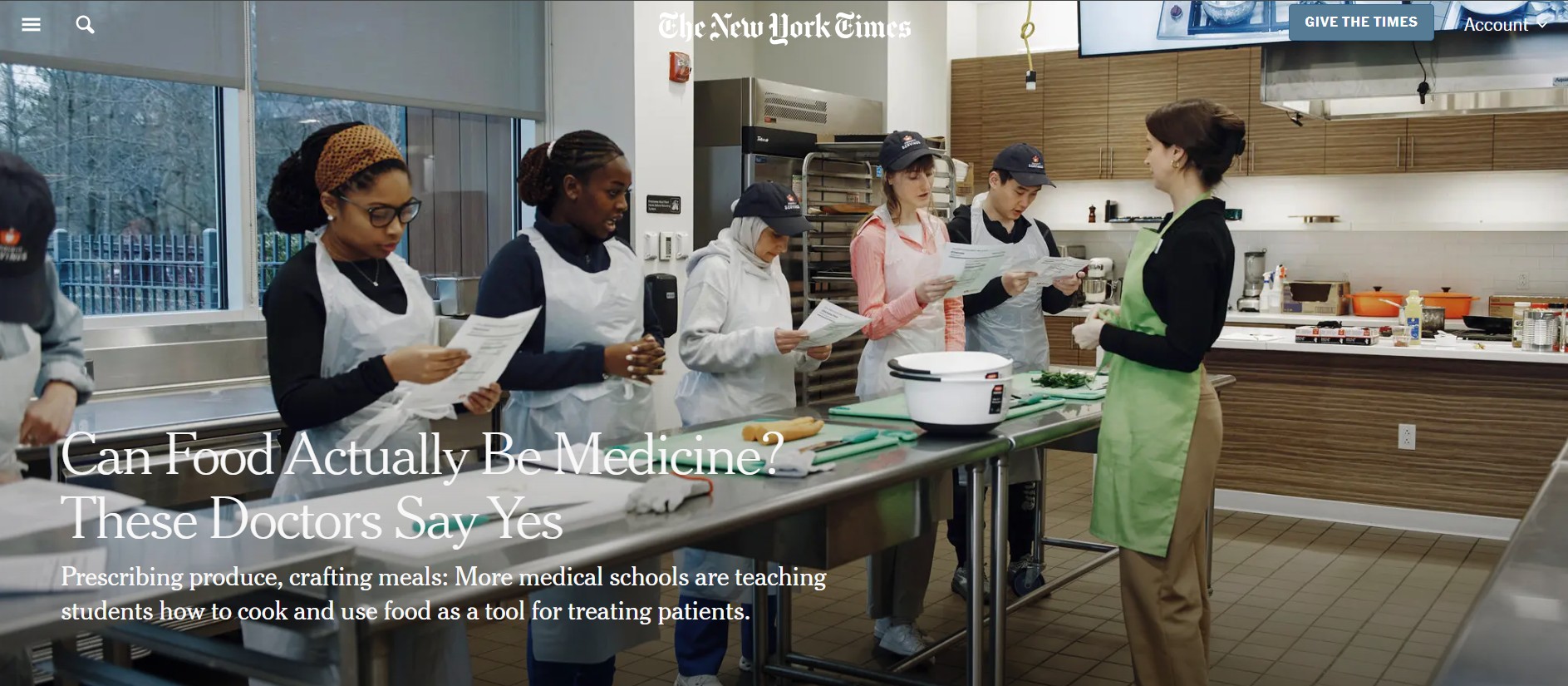 Medical students standing front of a metal workstation in a teaching kitchen. A woman in a bright green apron leads the class.
